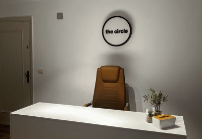 Modern reception area with a white front desk, tan leather chair, and circular wall branding.