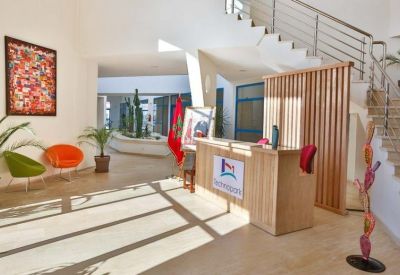 Bright sunlit lobby featuring a wooden reception desk, colorful lounge chairs, and a staircase.