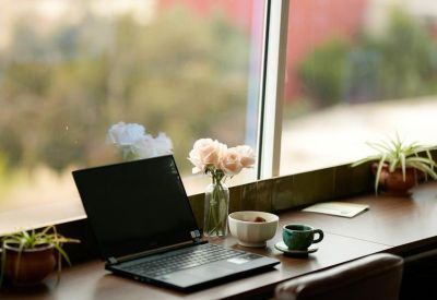 Laptop on a wooden window-side counter with coffee and flowers.