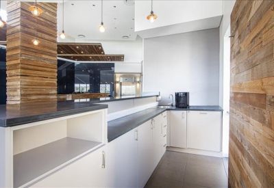 Modern office breakroom with white cabinetry and dark countertops beside a textured wood wall.