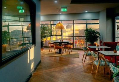 Sunlit communal lounge with warm wooden floors, potted plants, and a variety of seating areas.