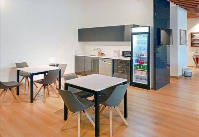 Sleek communal kitchen area with wood-style flooring, dining tables, and a glass-door fridge.