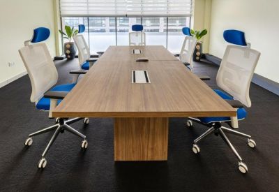 Bright conference room with a long wood-grain table and several ergonomic white and blue chairs.