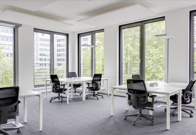 Bright open-plan workspace with multiple white desks and ergonomic black chairs near large windows.