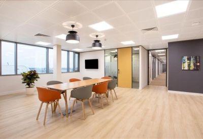 Bright coworking area with a long wooden table, orange and grey chairs, and large windows.