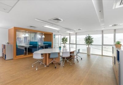 Lounge area with a large oval table, blue upholstered booths, and tall potted plants.