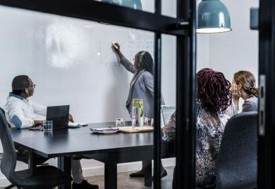 Team meeting in progress with a person writing on a whiteboard in a glass-walled conference room.