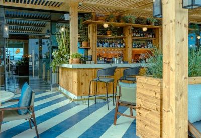 Sunlit communal lounge and bar area with wooden finishes, striped flooring, and outdoor seating.