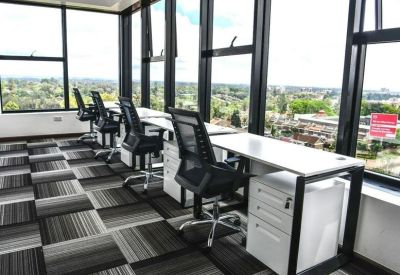 Row of modern workstations with white desks and black ergonomic chairs facing large windows.