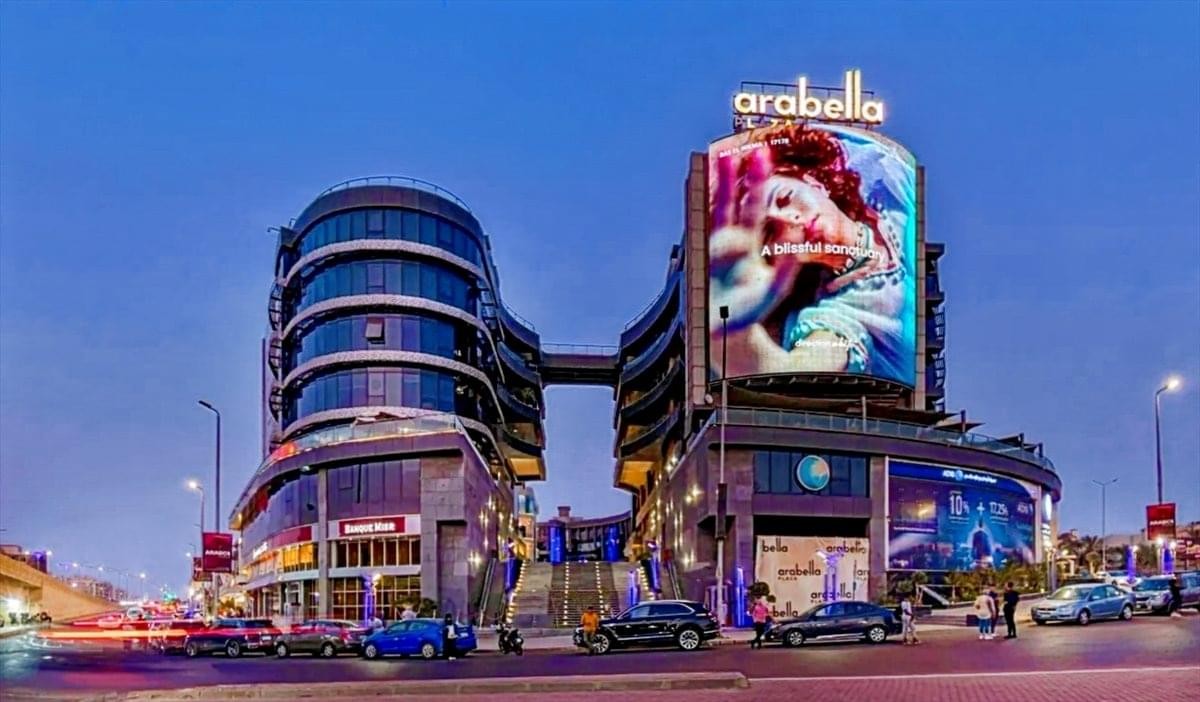 Dusk exterior of Arabella Plaza featuring illuminated digital billboards and modern walkways.