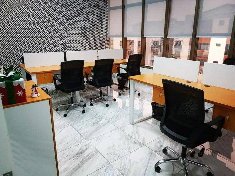 Open-plan office with multiple rows of wooden desks and large windows with blinds.