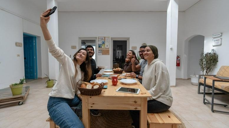 A group of people sitting around a long wooden table taking a selfie in a bright common area.